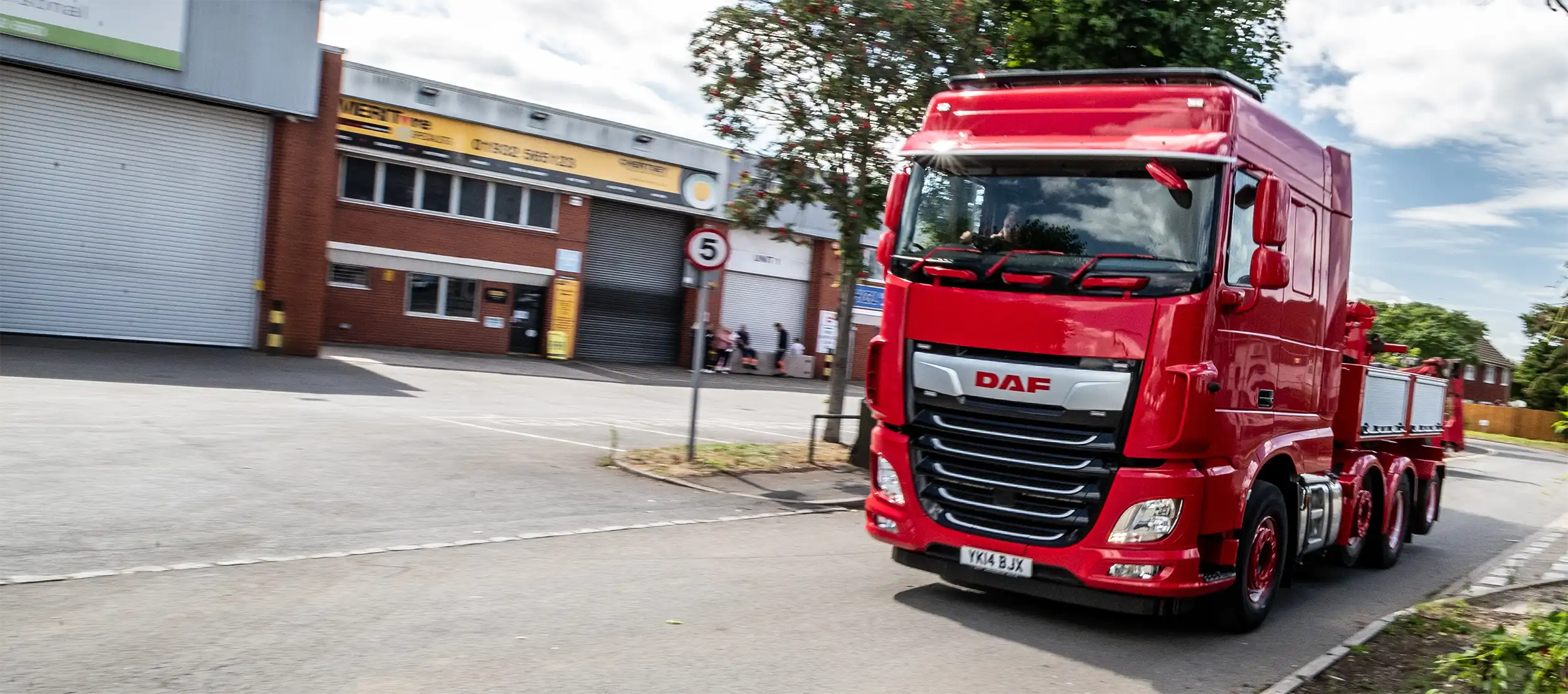 Red DAF heavy-duty truck driving on a road near brick industrial buildings and trees.