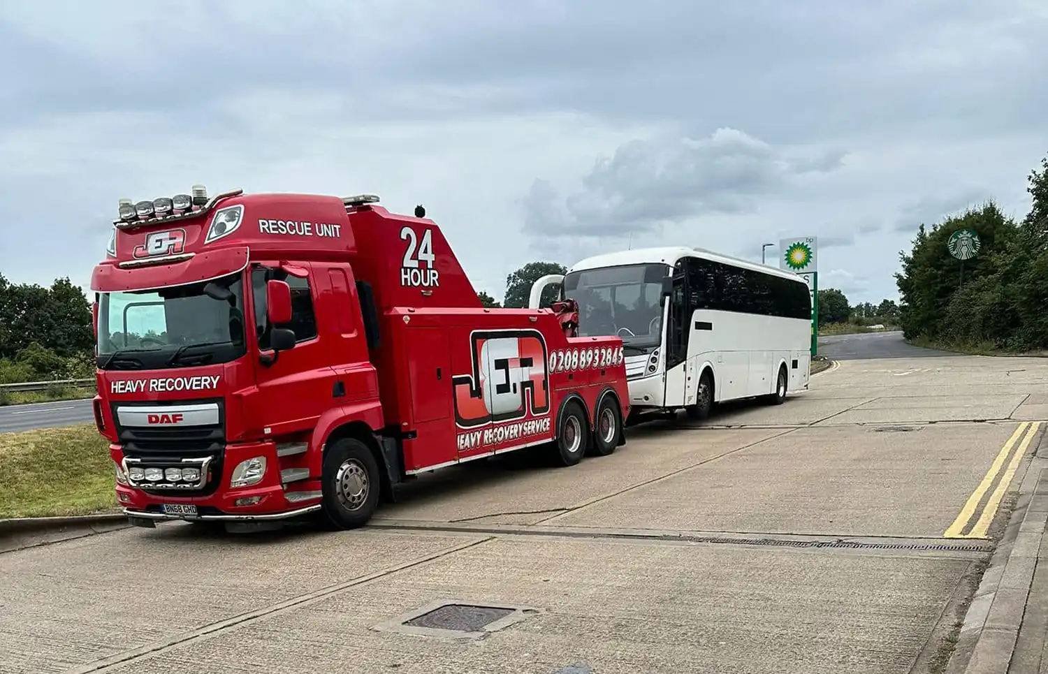 Red heavy recovery tow truck towing a white passenger bus on a roadside.