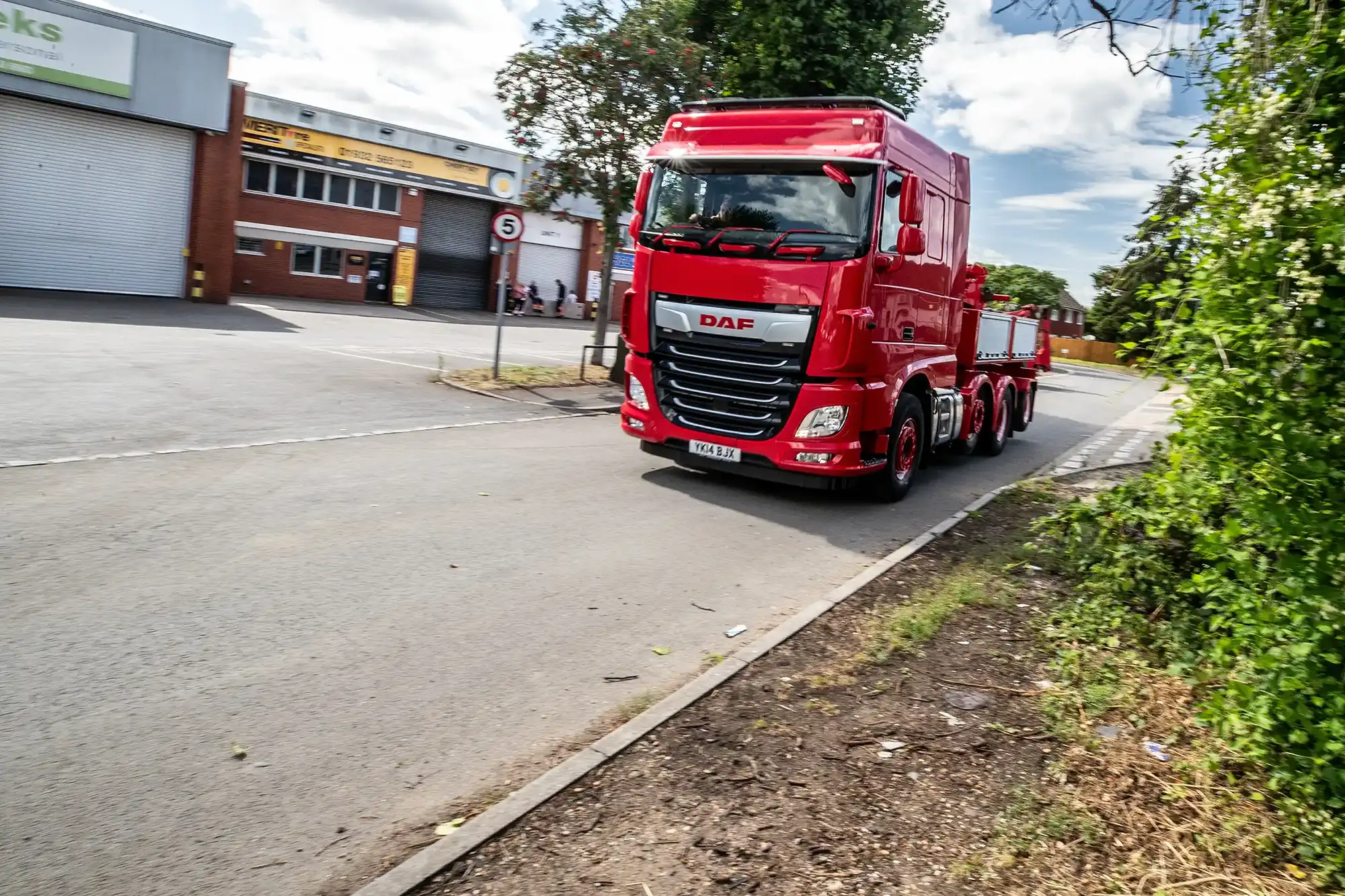 Red DAF truck driving on a street near industrial buildings and greenery on a partly cloudy day.