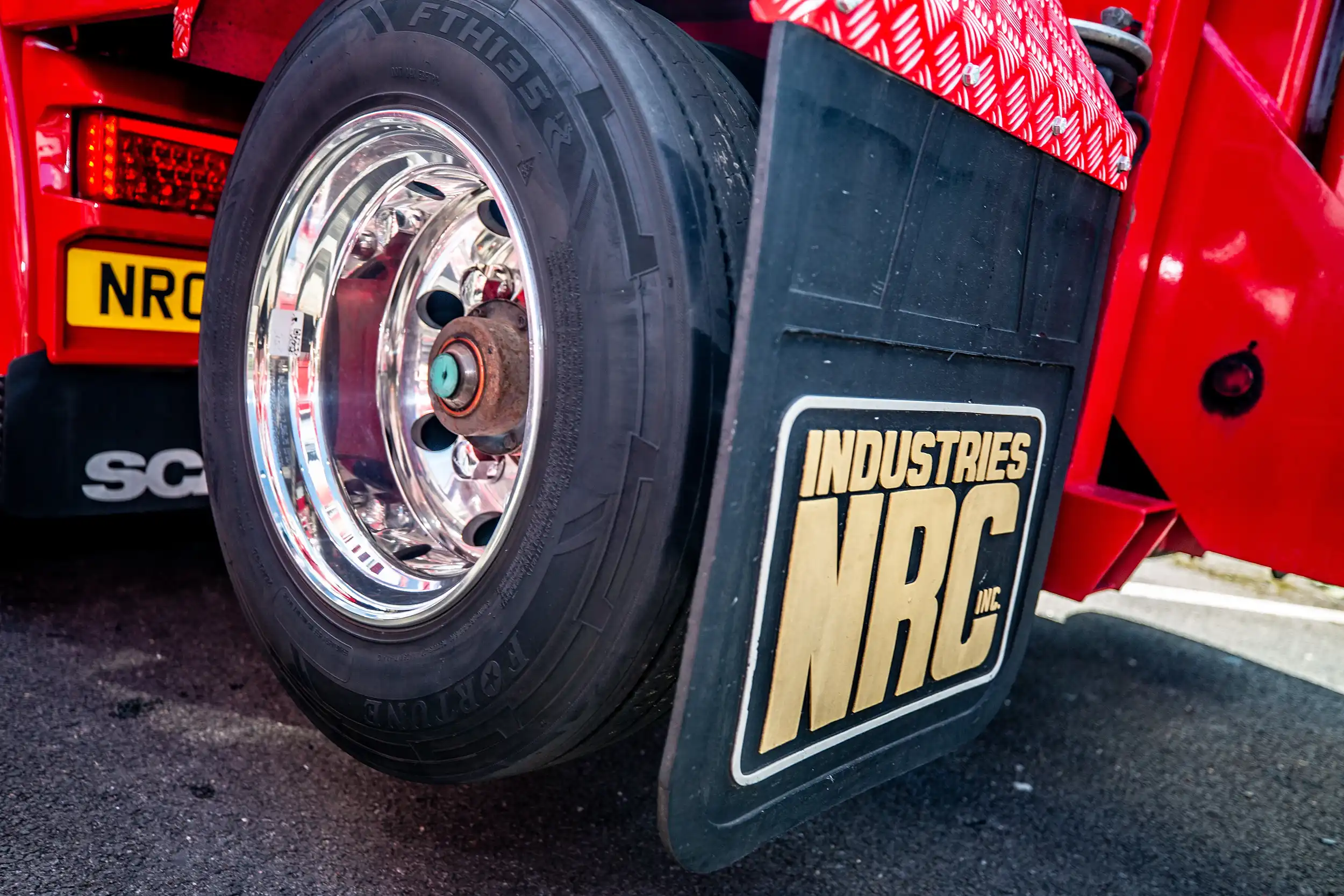 Close-up of a truck's shiny wheel and black mud flap with gold INDUSTRIES NRC logo on red body.