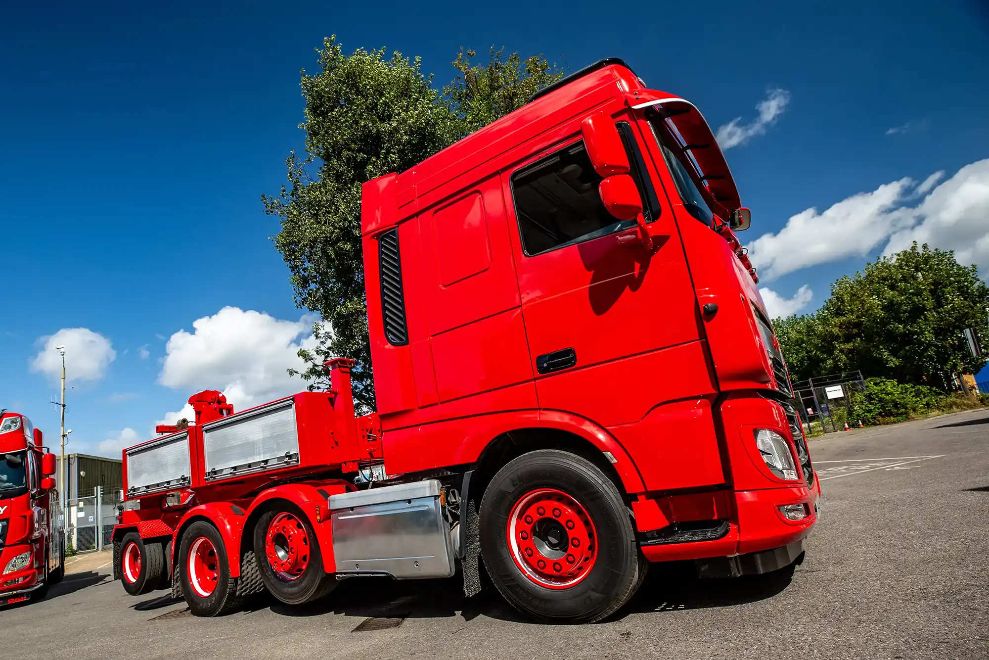 Side view of a bright red semi-truck cab parked on asphalt with blue sky and greenery behind.