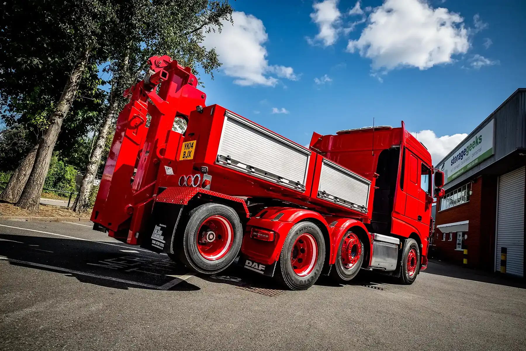 Red heavy-duty recovery truck parked outdoors under blue sky with trees and a building in background.