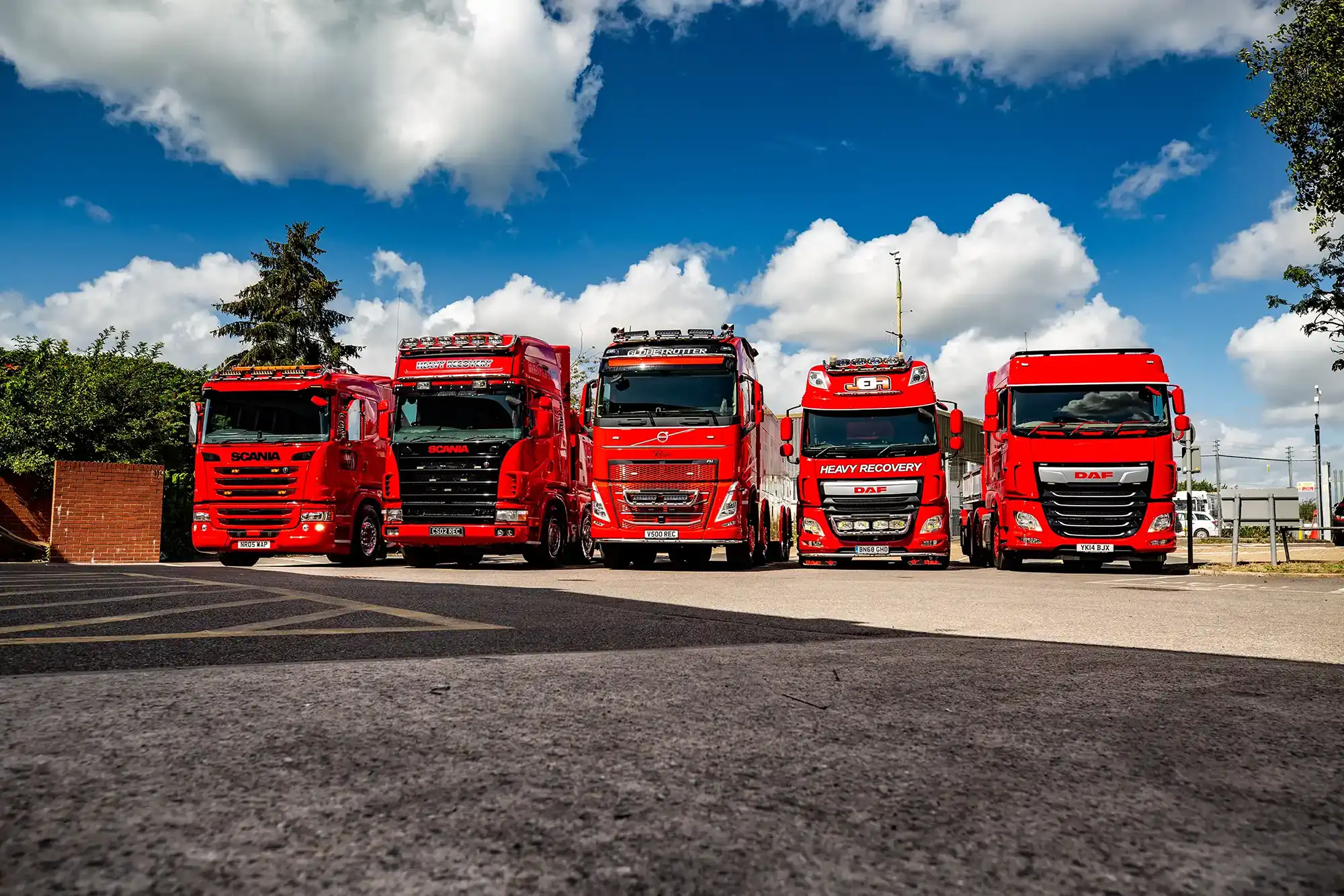 Five red heavy recovery trucks parked side by side under a blue sky with white clouds.