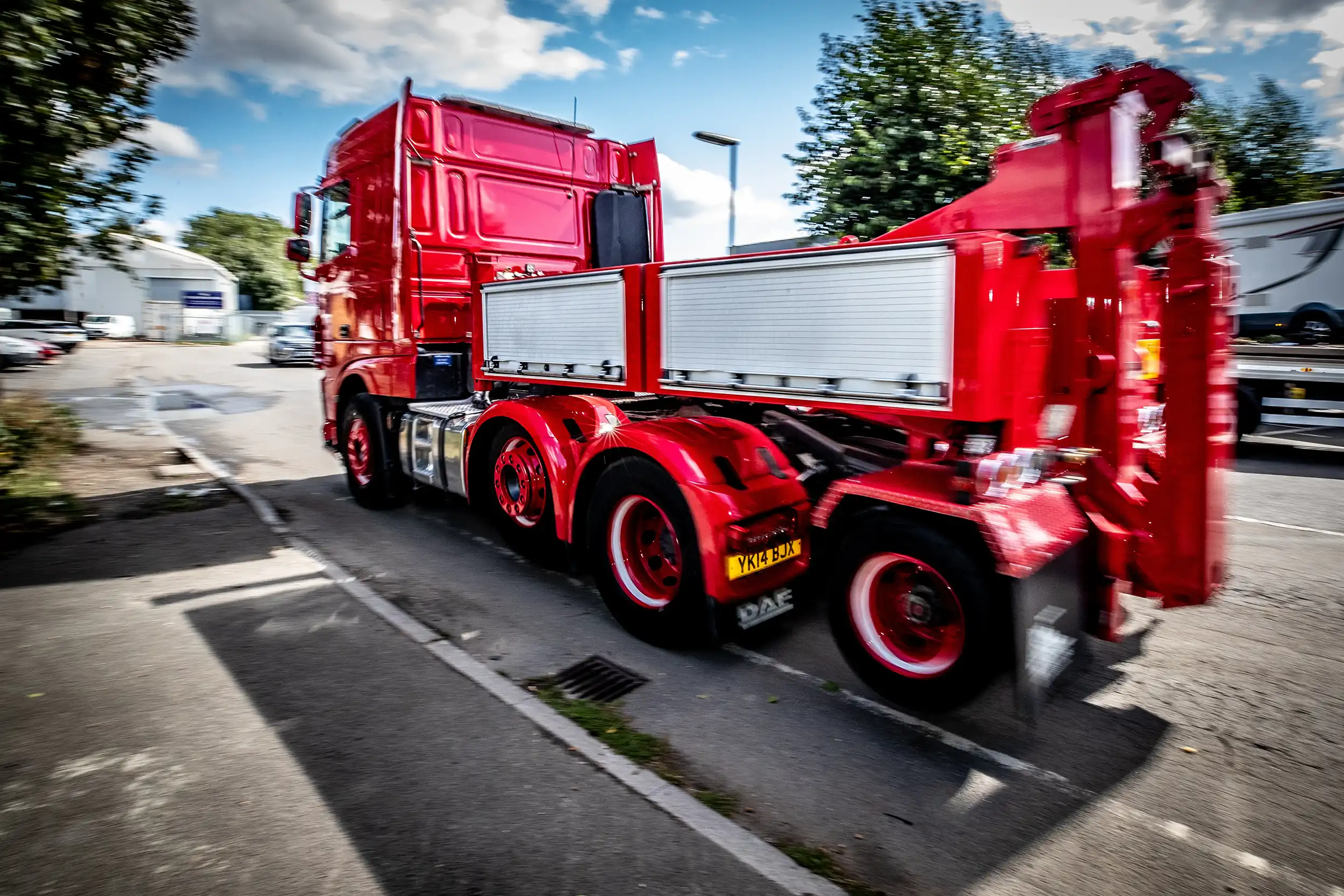 Red heavy-duty truck with flatbed trailer parked on roadside under a partly cloudy sky.