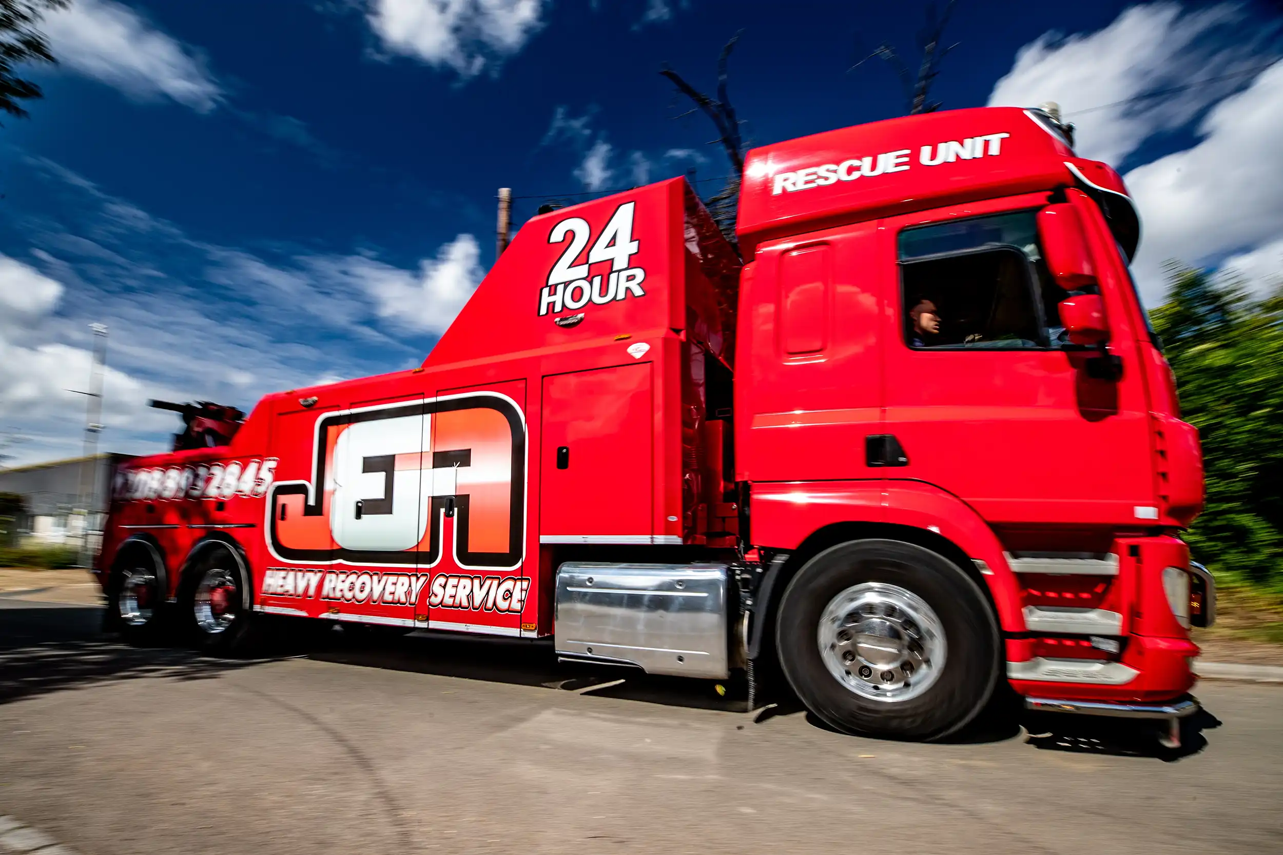 Red heavy recovery truck with 'JFA 24 Hour Rescue Unit' driving on road under blue sky.