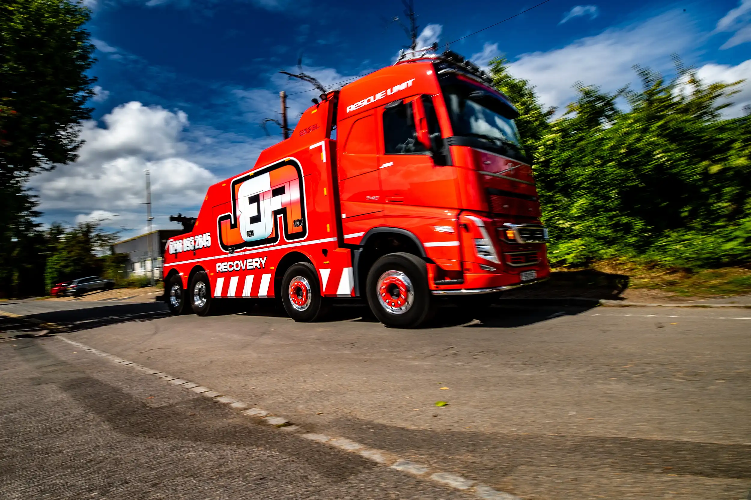 Red heavy-duty recovery truck driving on a road with trees and blue sky in the background.
