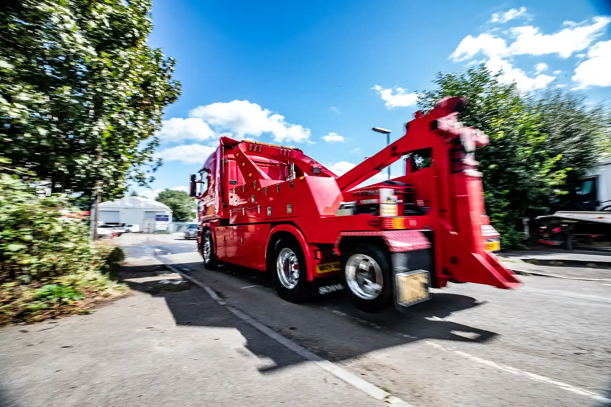 Red tow truck driving on a sunlit street with trees and buildings in the background.