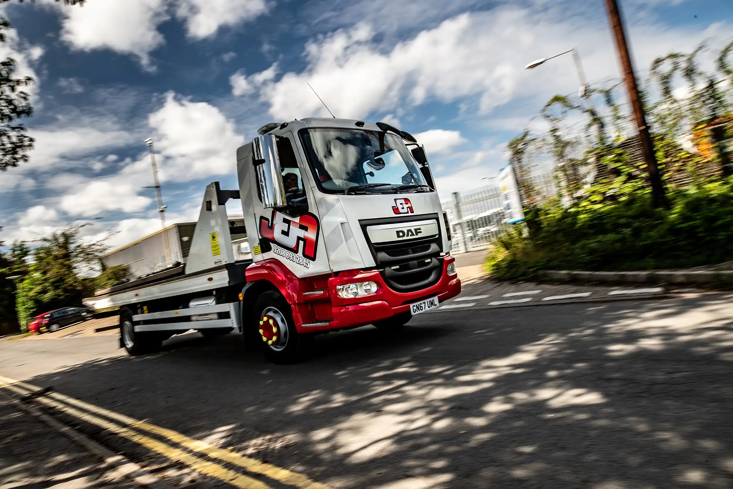 White and red flatbed tow truck with J&A logo driving on a road under a partly cloudy sky.