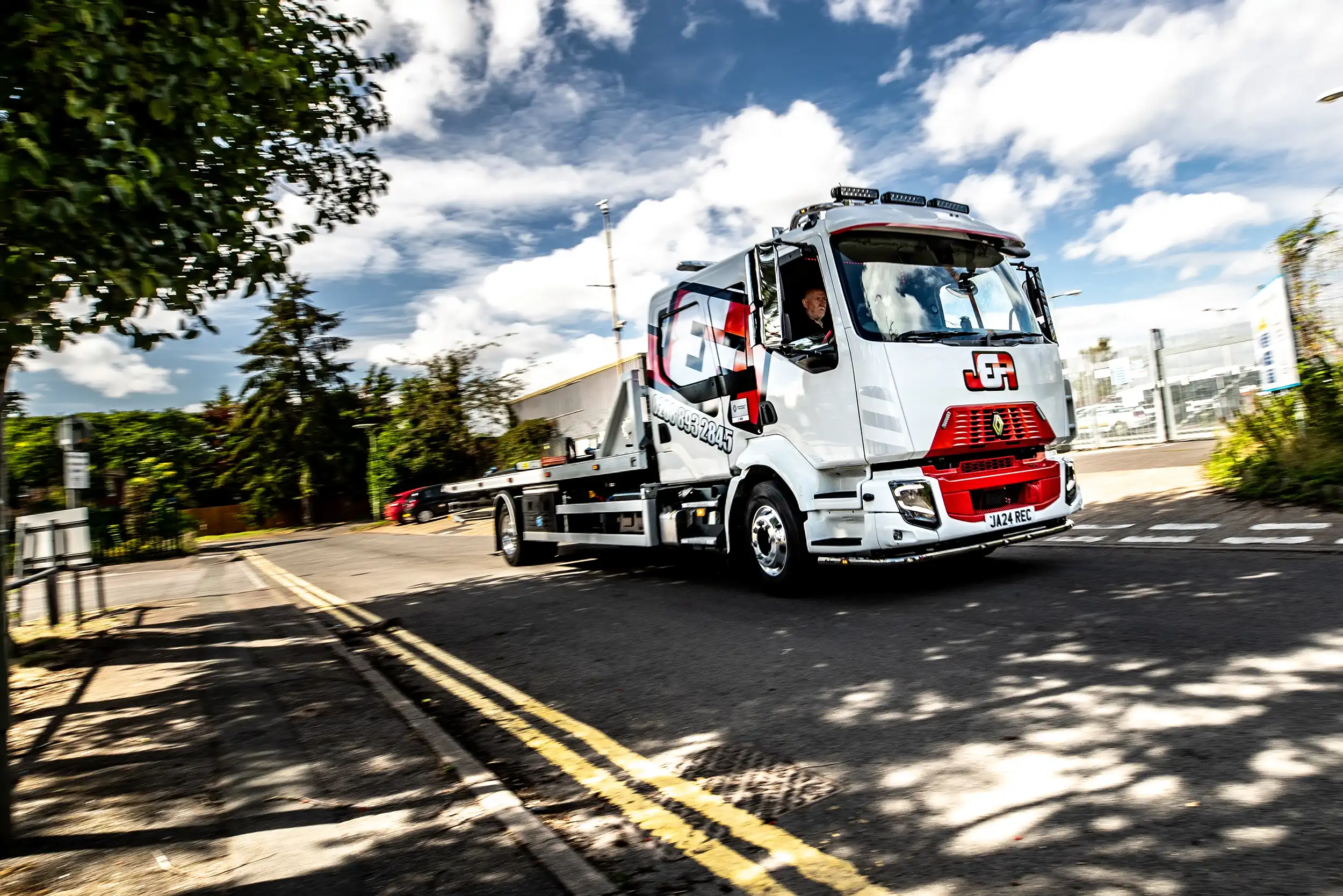 White flatbed tow truck driving on a sunny road with a blue sky and scattered clouds.