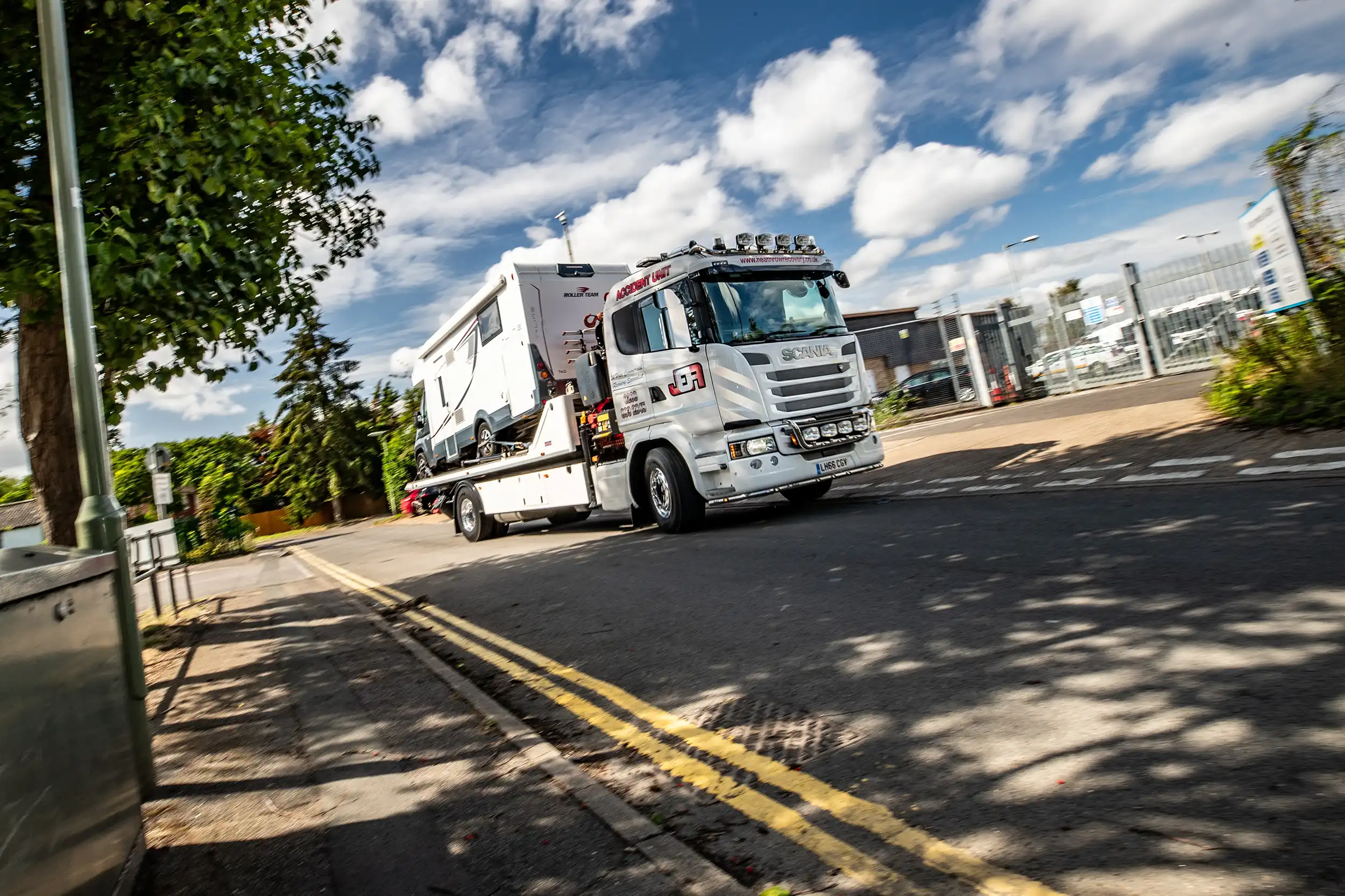 Tow truck carrying a white camper van driving on a street under a blue sky with clouds.