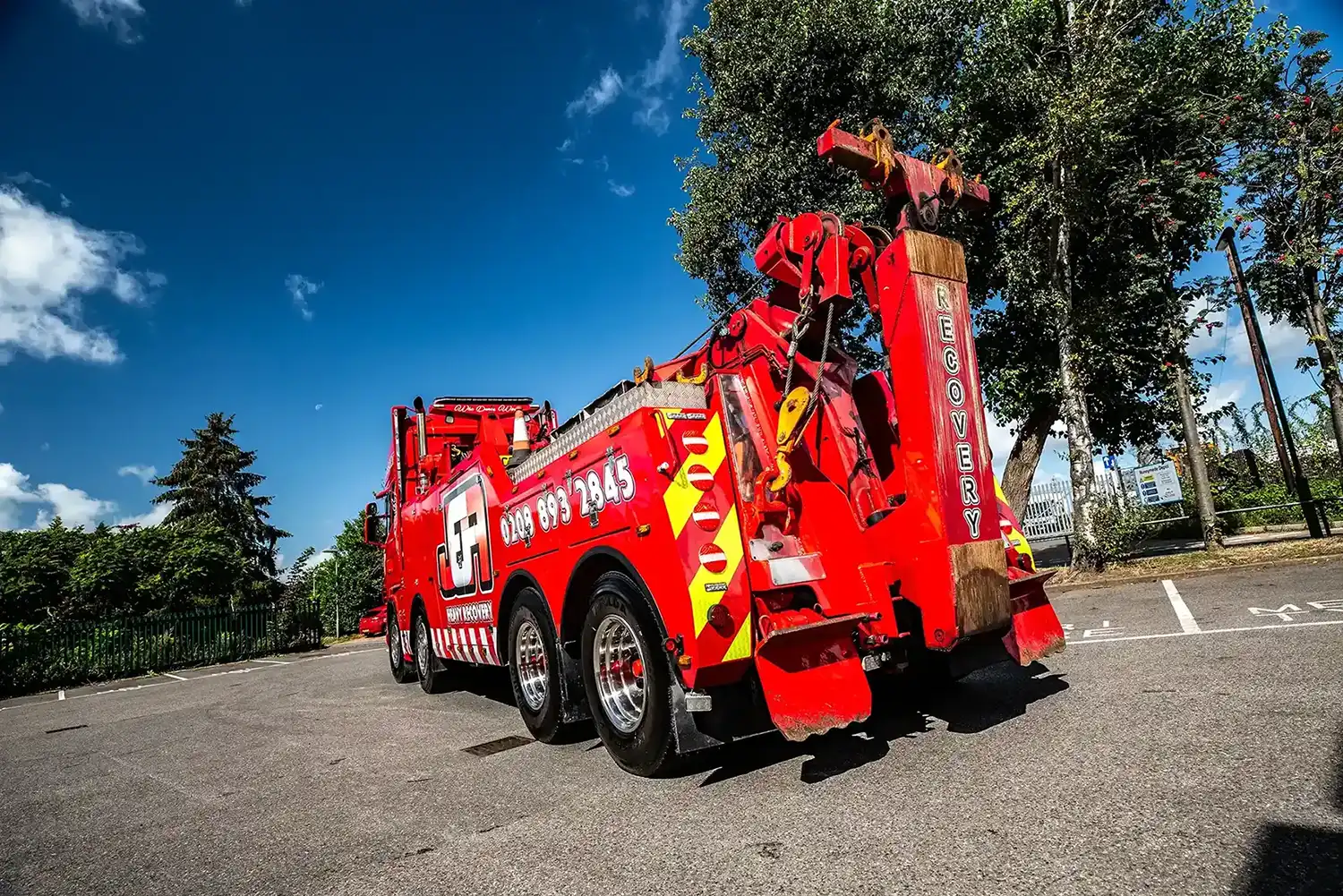 Red heavy recovery tow truck parked on road under clear blue sky near trees and fence.