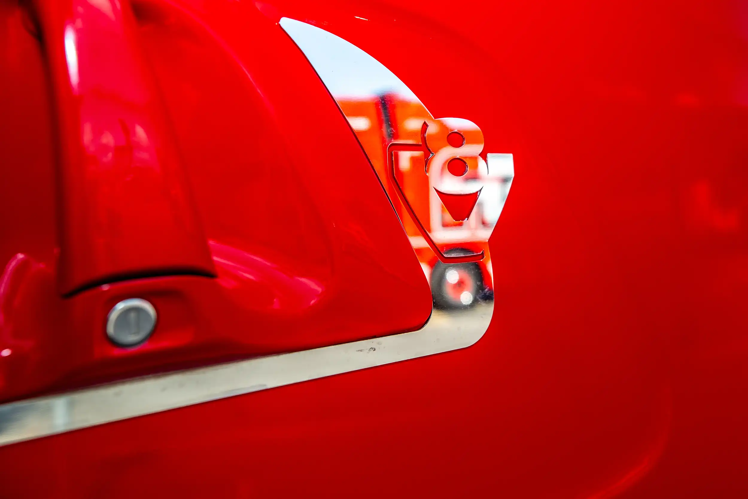 Close-up of a silver V8 emblem on a glossy red vehicle surface with part of a red side mirror.