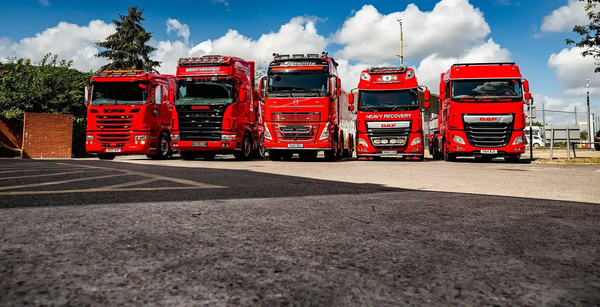 Five red heavy-duty recovery trucks lined up side by side under a blue sky with clouds.
