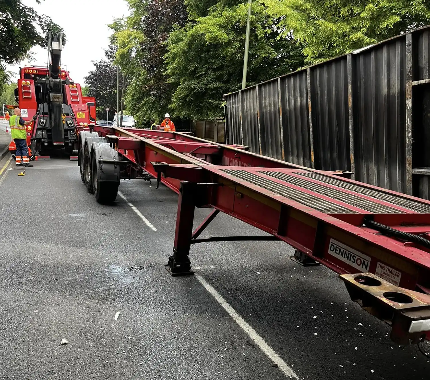 Red trailer chassis on a road with two workers in safety gear and a red tow truck in the background.