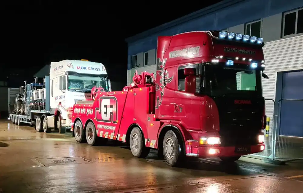 Red heavy recovery truck towing a white truck loaded with vehicles at night outside a building.